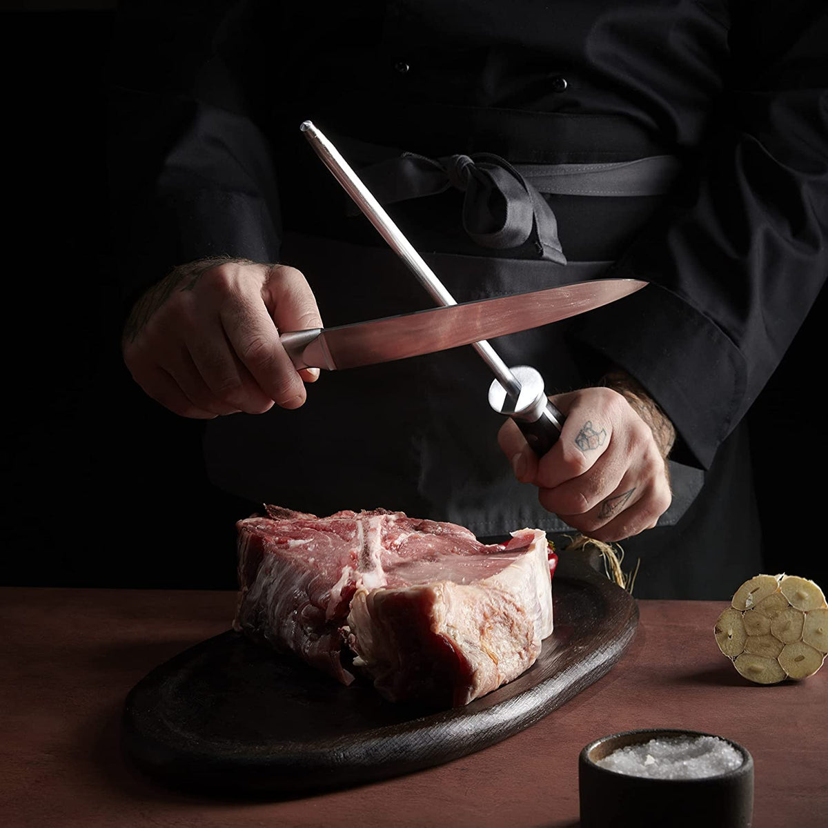 Product image 7 of 8: Chef sharpening kitchen knife with honing steel over raw steak, ready for precise cutting
