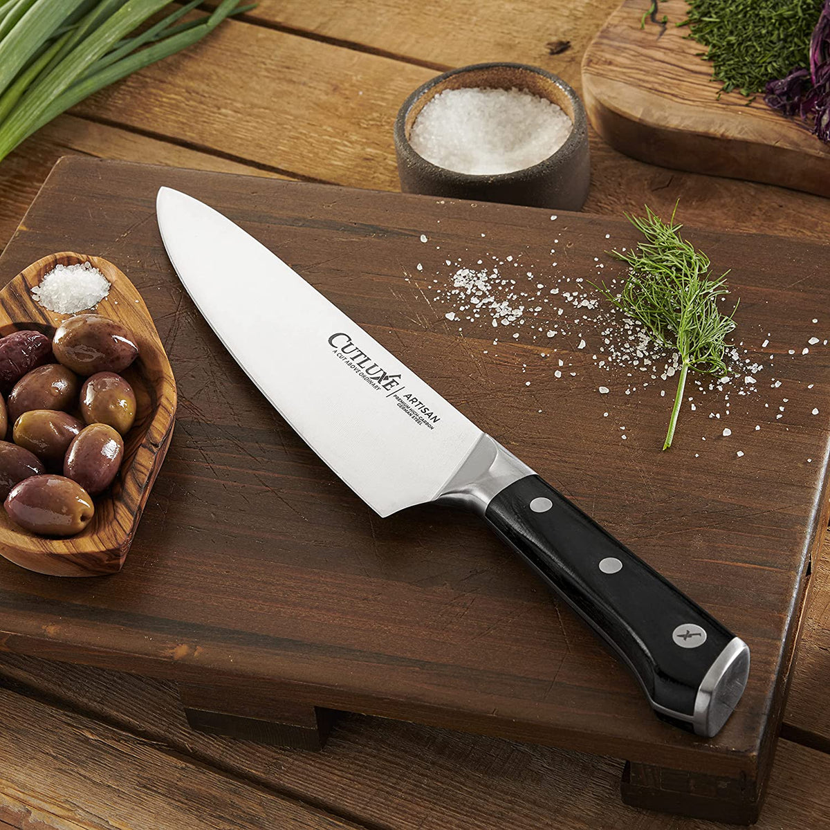 Product image 7 of 9: Cutluxe Artisan chef knife on wooden cutting board with olives, salt, and herbs for home cooking