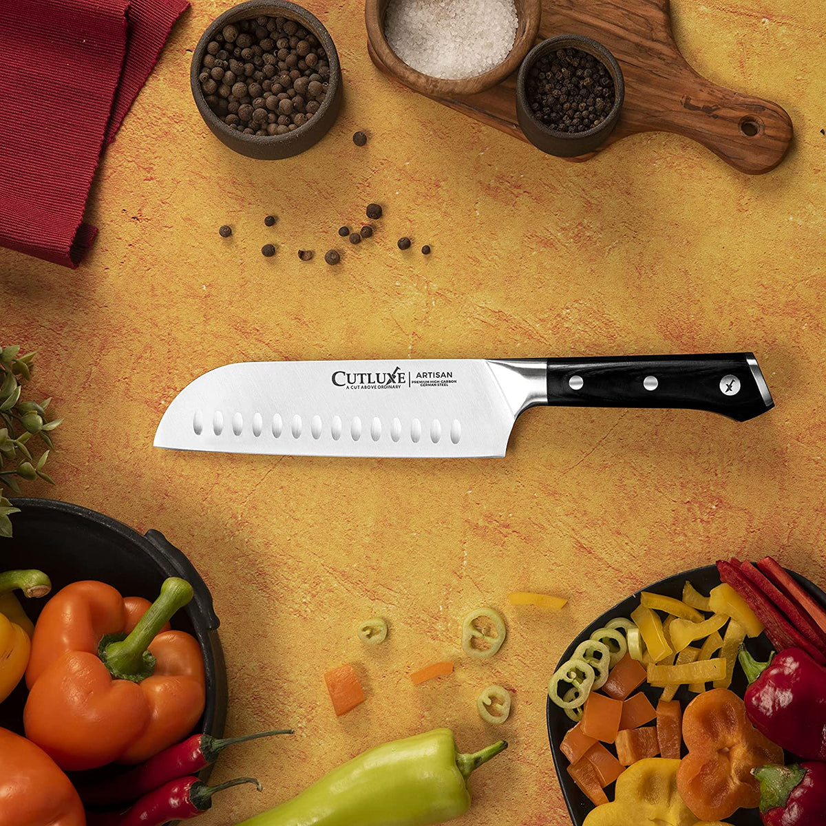 Product image 2 of 8: Cutluxe Artisan santoku knife with black handle, surrounded by fresh peppers, salt, and peppercorns on a warm countertop