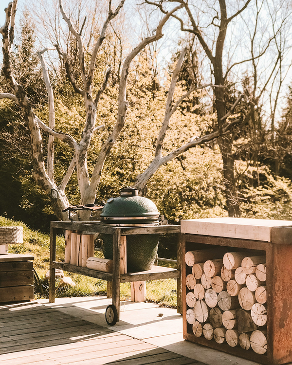 Green ceramic kamado grill on wooden cart beside firewood stack in sunny backyard