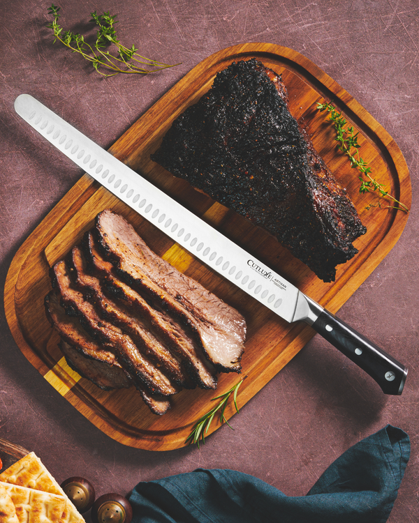 Sliced smoked brisket on wooden board with Cutluxe carving knife, fresh herbs, and bread